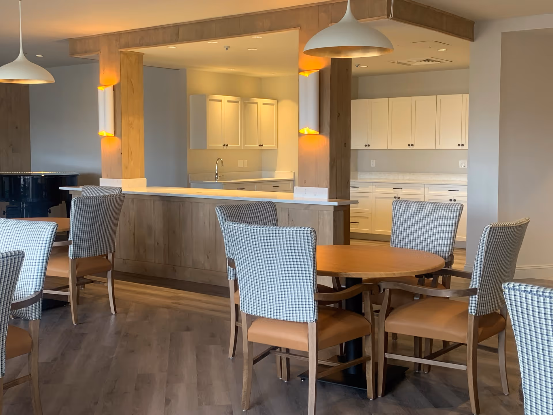 A dining area with round wooden tables and cushioned chairs featuring a checkered pattern. The space has wooden flooring and is adjacent to a kitchen area with white cabinets and countertops. Pendant lights hang from the ceiling, and a black grand piano is visible in the background.