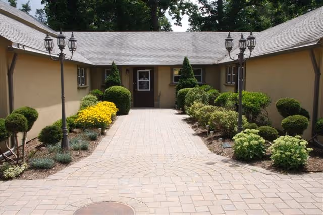 Paved walkway leading to the entrance of a single-story building with beige walls and a gray shingled roof, flanked by neatly trimmed bushes, yellow flowers, and vintage-style street lamps on both sides.