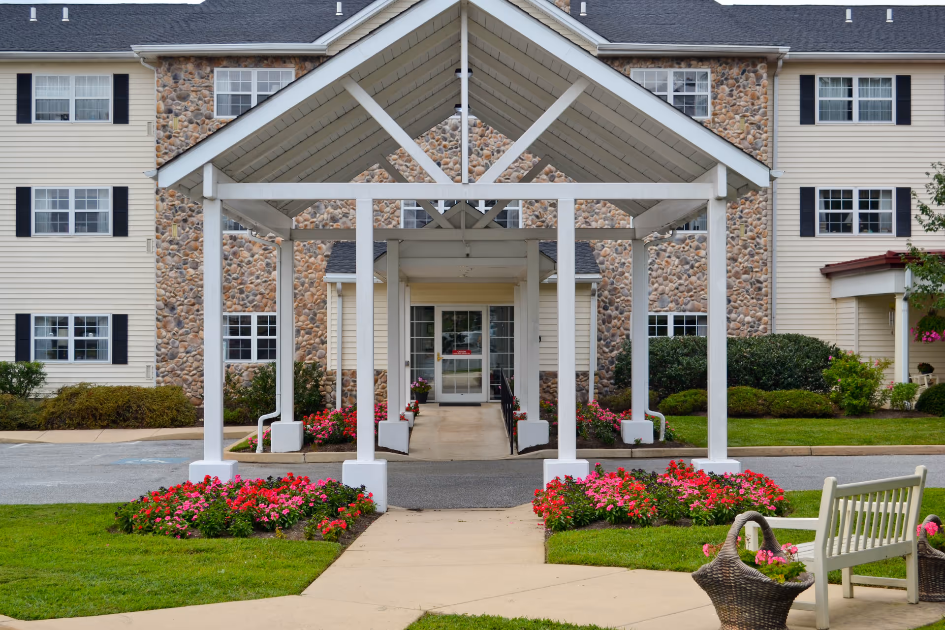 Entrance of a senior living facility with a covered walkway supported by white pillars, flower beds with pink flowers on either side, a stone and beige siding building in the background, and a white bench with a basket planter nearby.