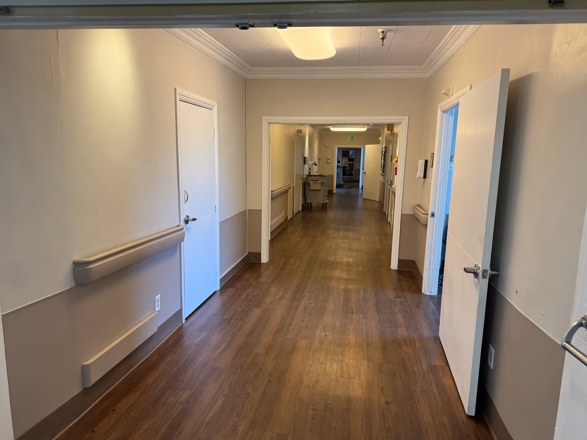 A long, empty hallway in a senior living facility with wood-look flooring, beige walls with handrails, and several closed and open white doors along the sides. The hallway is well-lit with ceiling lights and has a clean, simple appearance.