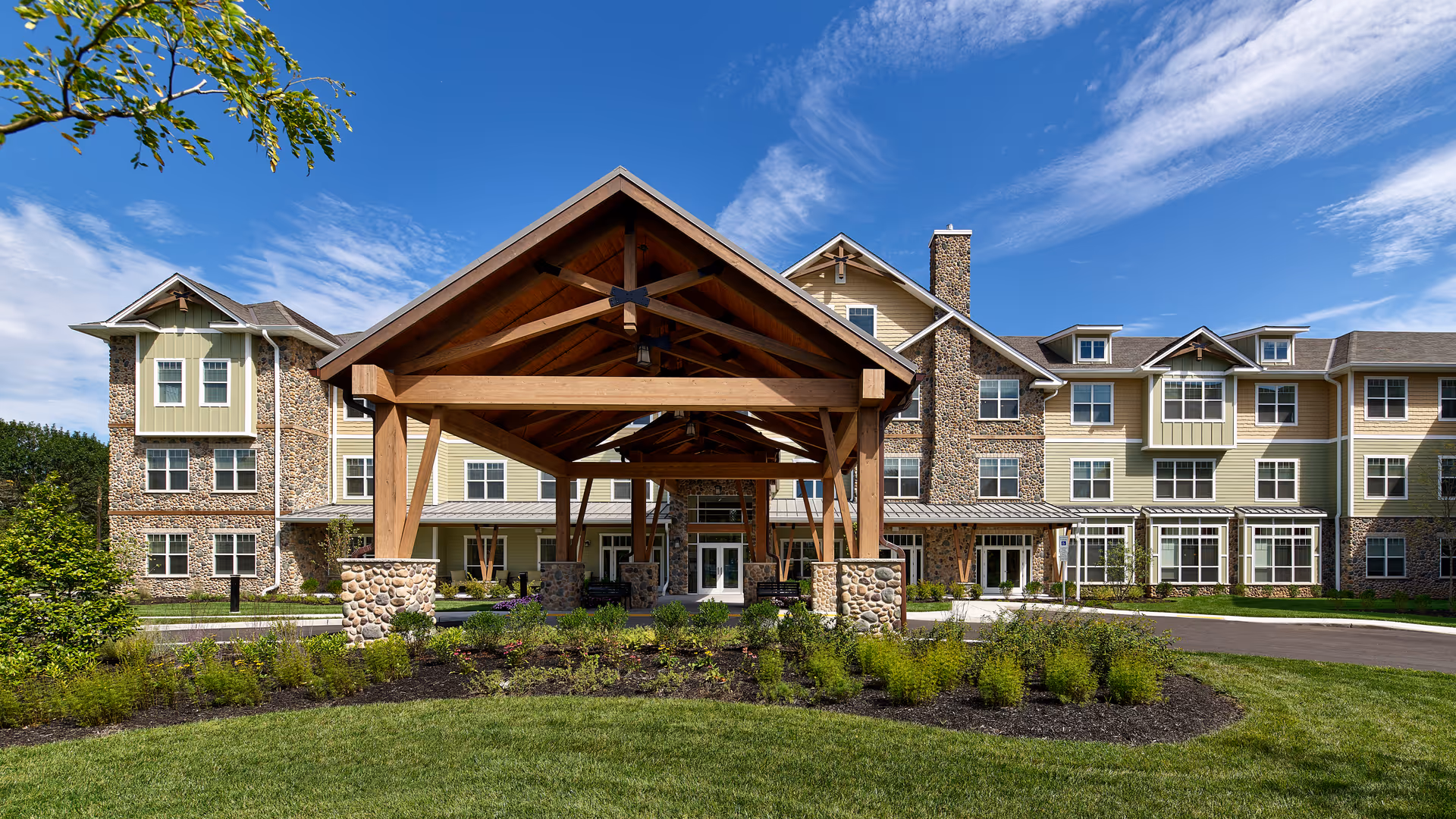 Front exterior view of The Chelsea at Greenburgh, a multi-story senior living facility with a large wooden porte-cochere supported by stone pillars, surrounded by landscaped greenery and a clear blue sky with some clouds.
