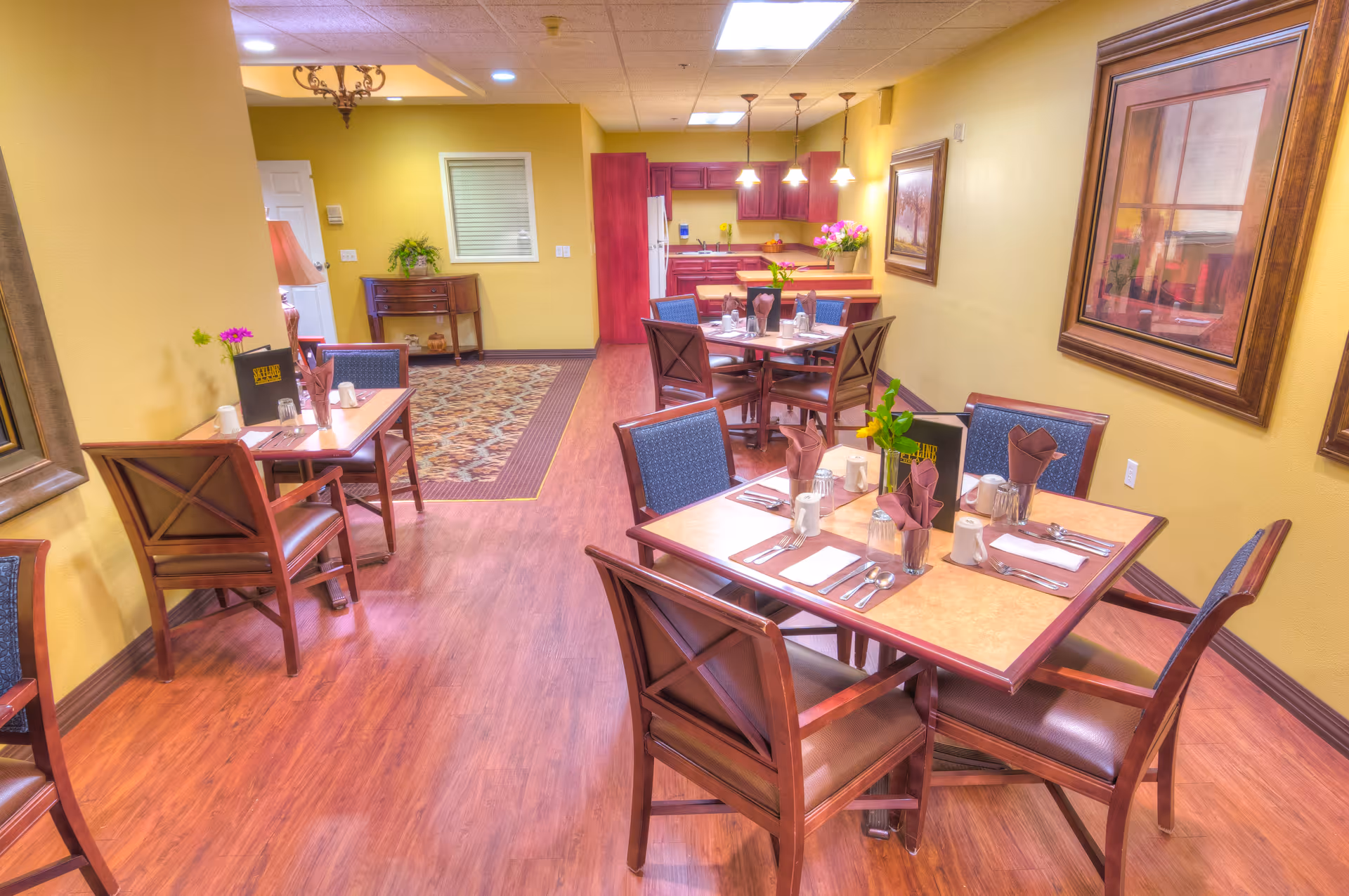 Interior view of a senior living facility dining room with several wooden tables and chairs. Each table is set with placemats, napkins, utensils, and small flower vases. The room has warm yellow walls, wooden flooring, framed artwork on the walls, and a kitchen area in the background with cabinets and hanging lights.