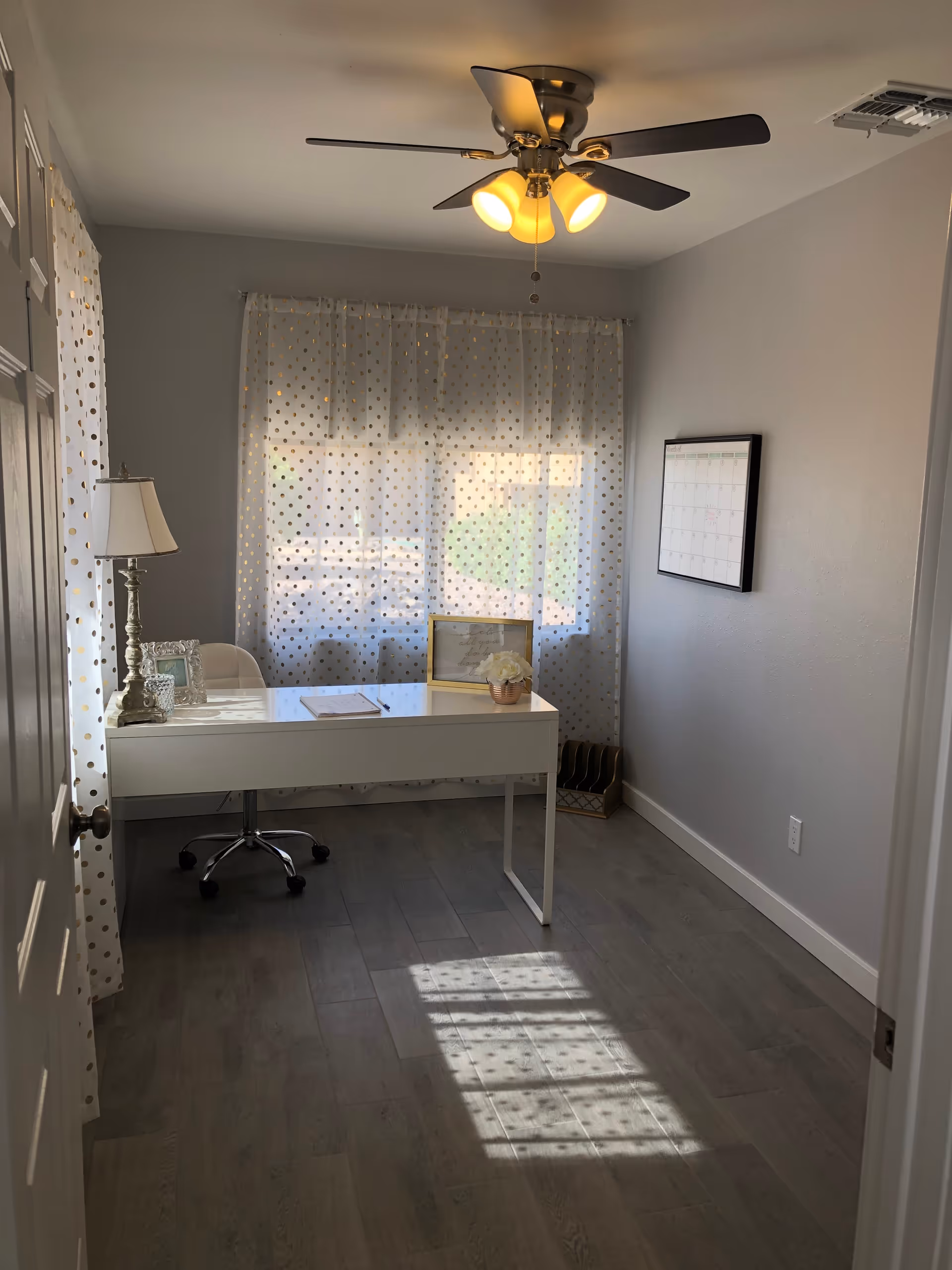 A small, bright office room with a white desk and a white swivel chair on wheels. The desk has a table lamp, a framed picture, a notebook, and a small flower pot. Sheer white curtains with gold polka dots cover the window, allowing sunlight to cast a patterned shadow on the wooden floor. A ceiling fan with lights is mounted on the ceiling, and a wall calendar is hung on the right wall.