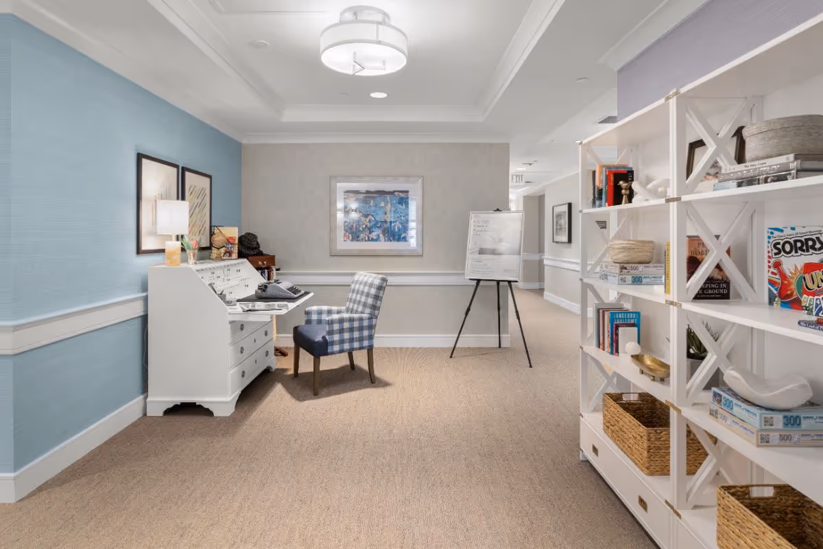 A well-lit hallway in a senior living facility with beige carpet and light blue and gray walls. On the left side, there is a white desk with a lamp, decorative items, and a checkered chair. On the right side, there is a white open shelving unit filled with books, board games, and baskets. A framed picture hangs on the far wall above a whiteboard on an easel.