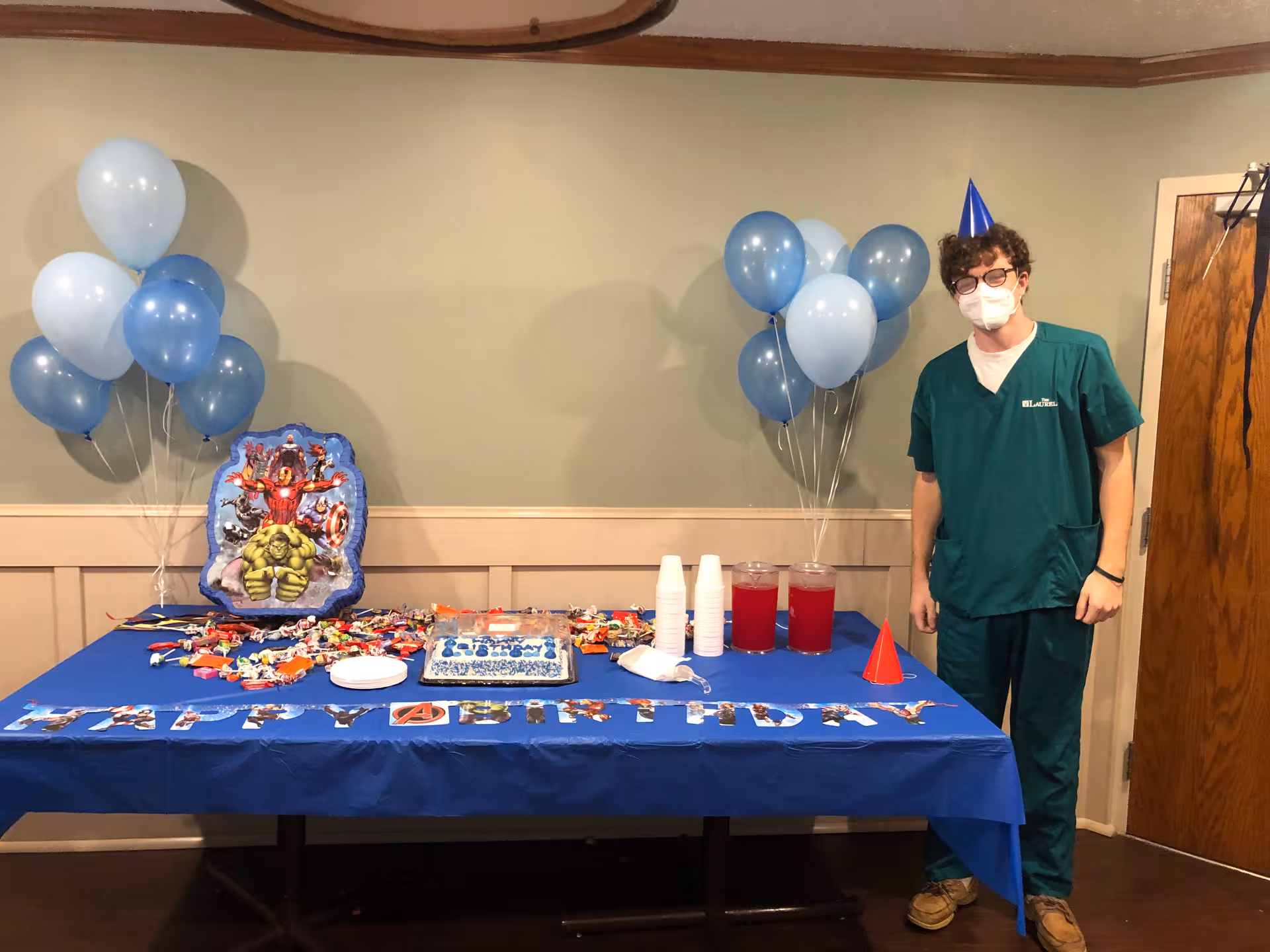 A person wearing green scrubs, a party hat, glasses, and a face mask stands next to a table decorated for a birthday party. The table has a blue tablecloth with 'HAPPY BIRTHDAY' spelled out in Avengers-themed letters, a birthday cake, two pitchers of red drink, disposable cups, assorted candies, and two bunches of blue and light blue balloons. The setting appears to be an indoor room with beige walls and a wooden door.
