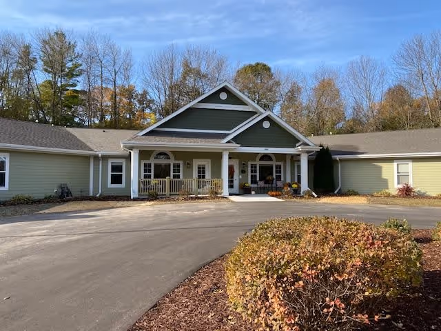 Front exterior view of a single-story building with green siding and a covered entrance with white columns. There is a paved driveway and landscaped bushes in front, with trees in the background under a blue sky.