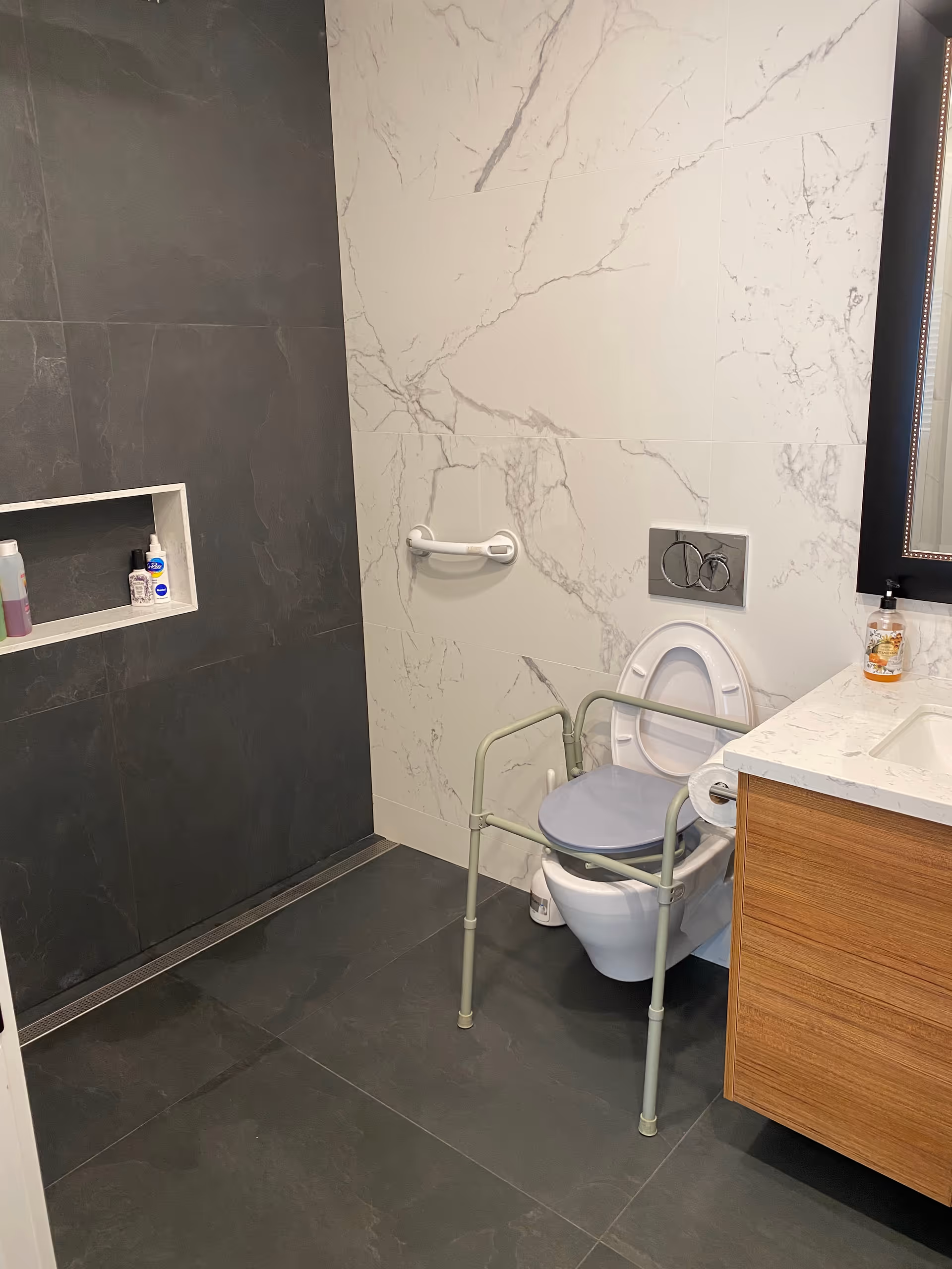 A modern bathroom with dark floor and wall tiles, a white marble-patterned wall behind a wall-mounted toilet equipped with a raised toilet seat and safety frame. There is a grab bar on the wall next to the toilet, a wooden vanity with a white countertop and a soap dispenser, and a recessed shelf in the shower area holding toiletries.
