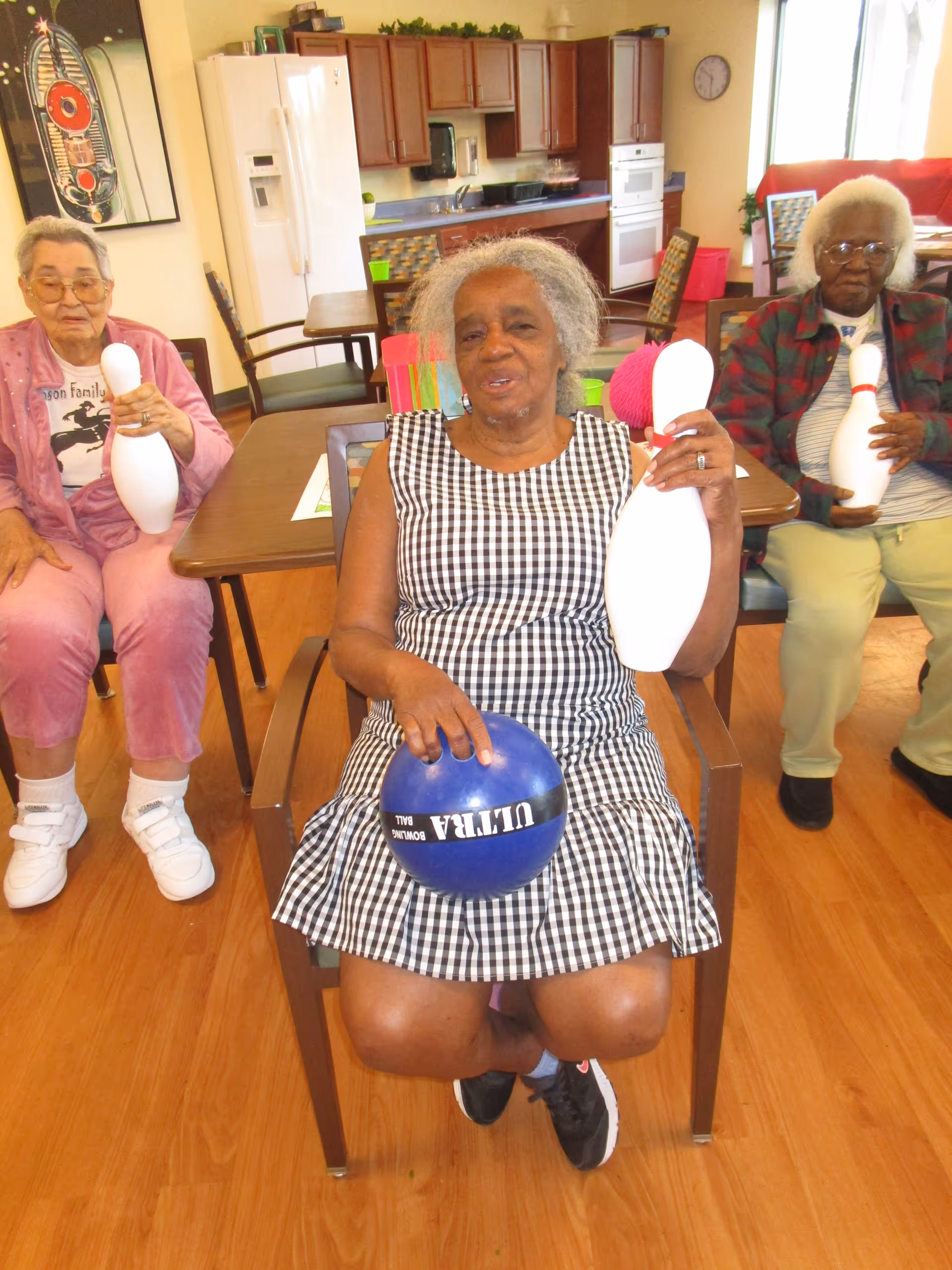 Three elderly women sitting in chairs inside a room with wooden floors and kitchen cabinets in the background. The woman in the center is holding a blue bowling ball and a white bowling pin, while the women on either side are each holding a white bowling pin. They appear to be participating in a recreational activity.