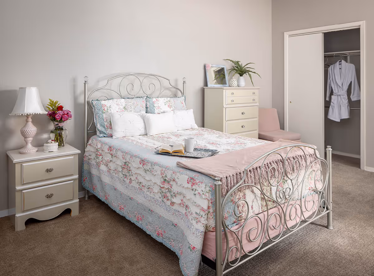 A cozy bedroom with a metal frame bed adorned with floral bedding and multiple pillows. On the bed is an open book and a white mug on a tray. To the left of the bed is a nightstand with a pink lamp, a small cake, and a vase of flowers. To the right is a cream-colored dresser with a plant and a framed picture on top, next to a pink chair and an open closet door revealing a white robe hanging inside. The room has beige carpet and light-colored walls.