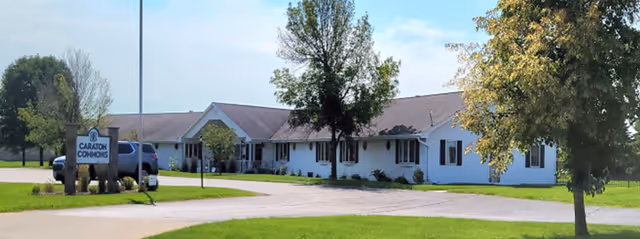 Single-story white building with a brown roof surrounded by green grass and trees under a partly cloudy sky. A sign near the driveway reads 'Caraton Commons'. Several vehicles are parked near the building.