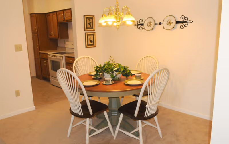 A dining area with a round wooden table set with four plates and cups, surrounded by four white wooden chairs with dark seats. A decorative plant centerpiece is on the table. The room has beige carpet and white walls, with a brass chandelier hanging above the table. In the background, a kitchen with wooden cabinets and a white stove is visible. Wall decorations include two framed pictures and a decorative plate holder with two plates.