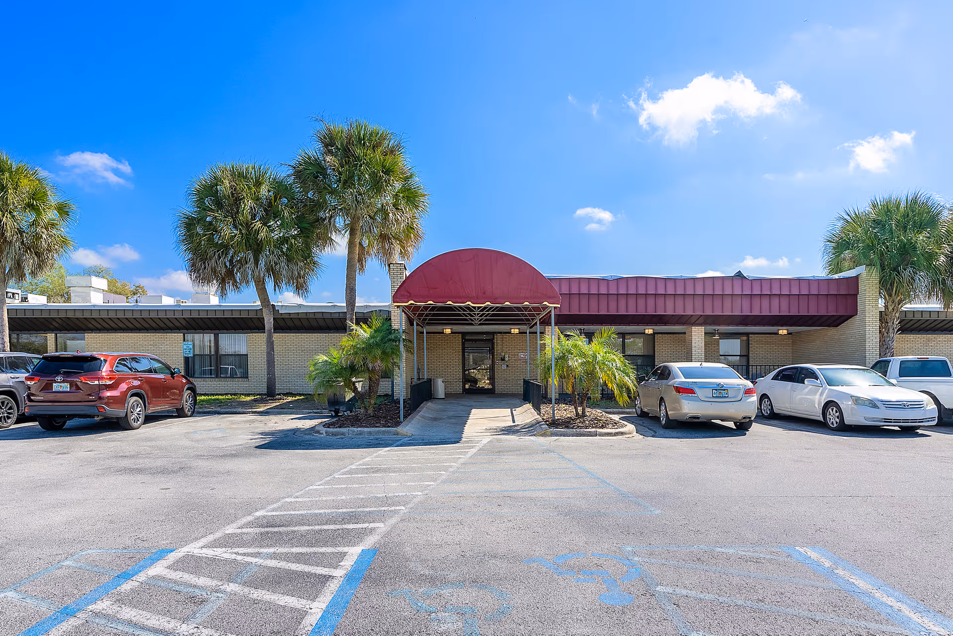 Front exterior view of Haines City Rehabilitation and Nursing Center showing a single-story building with a maroon awning over the entrance. Several palm trees and parked cars are visible in the parking lot under a blue sky with some clouds.