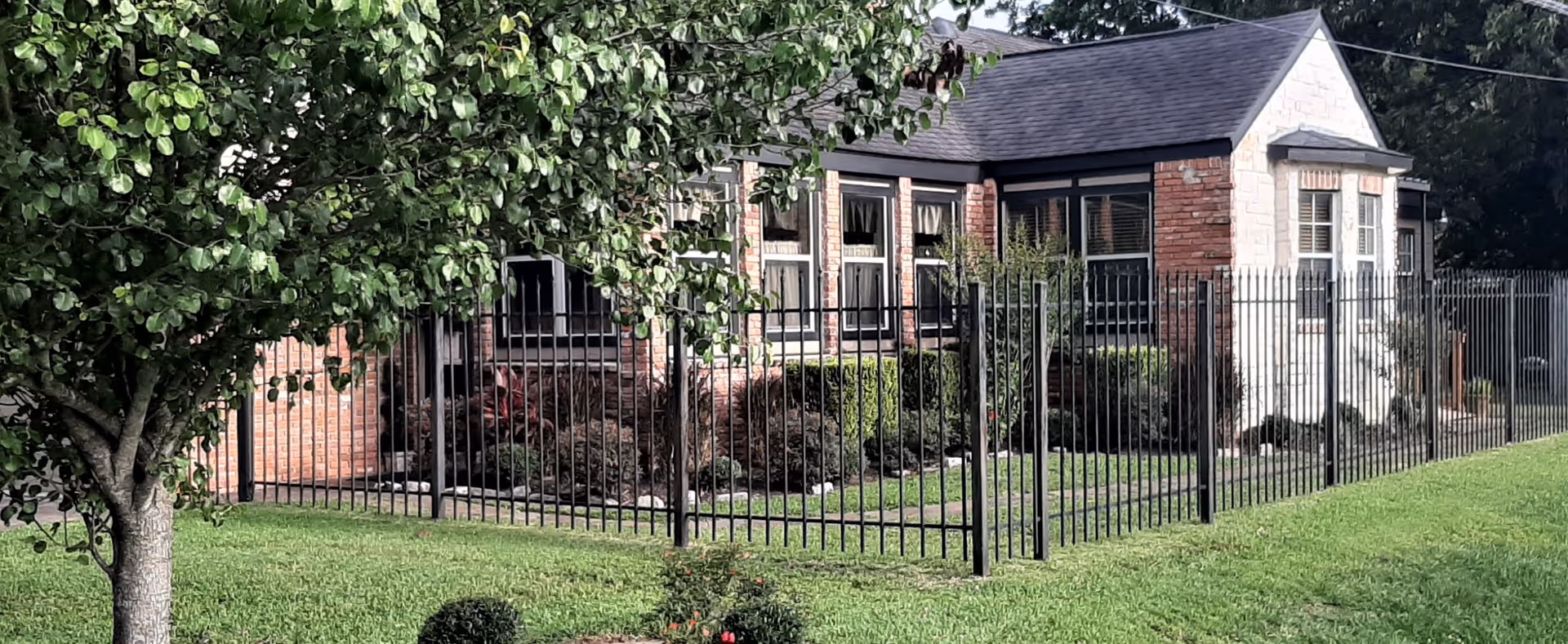 Exterior view of a brick and stone building surrounded by a black metal fence with a green lawn and a tree in the foreground.
