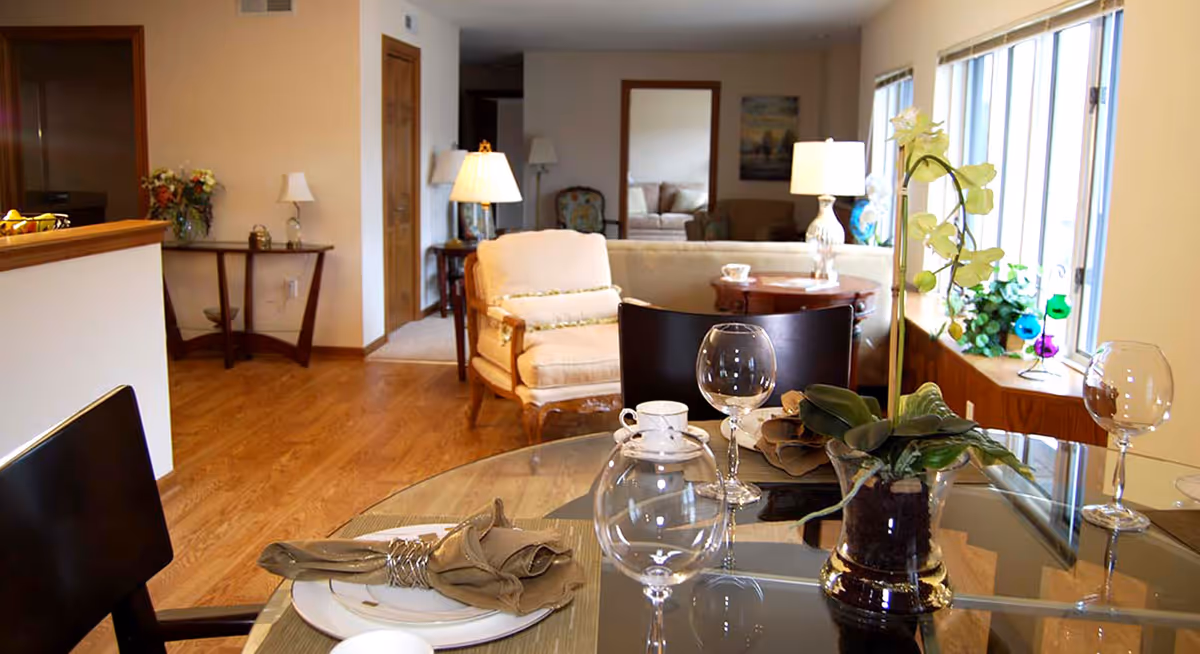 A cozy living and dining area in an independent living facility. The foreground shows a glass dining table set with plates, napkins, cups, and wine glasses. A small potted plant is on the table. In the background, there is a beige armchair, a sofa, side tables with lamps, and large windows letting in natural light. The room has wooden flooring and light-colored walls.