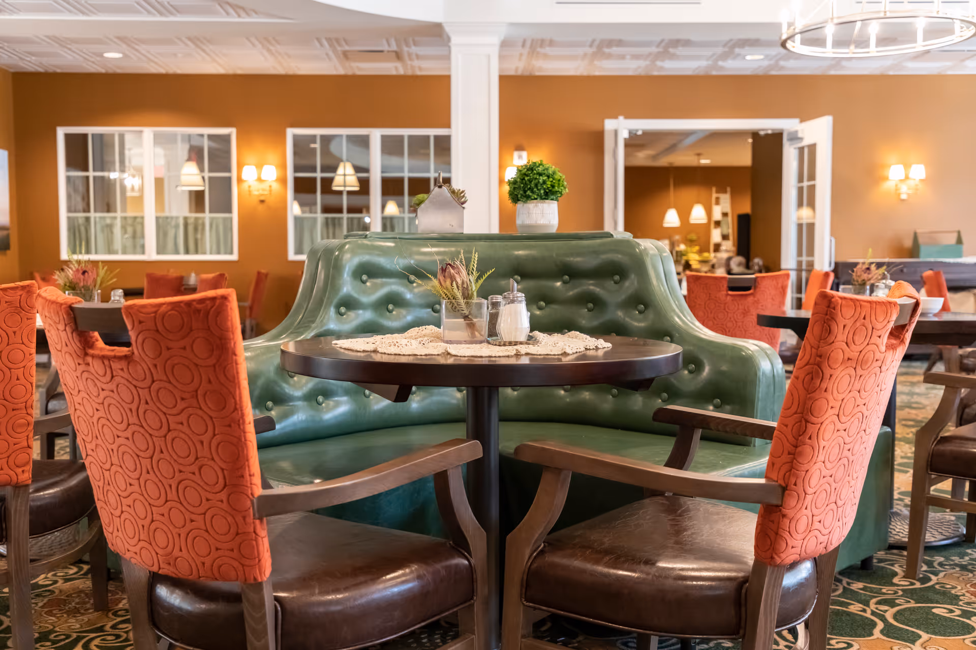 A cozy dining area with a round dark wooden table surrounded by a green tufted leather booth and four orange upholstered chairs with circular patterns. The room has warm brown walls, white ceiling tiles, and wall sconces providing soft lighting. In the background, there are windows and an open doorway leading to another room with more tables and chairs.