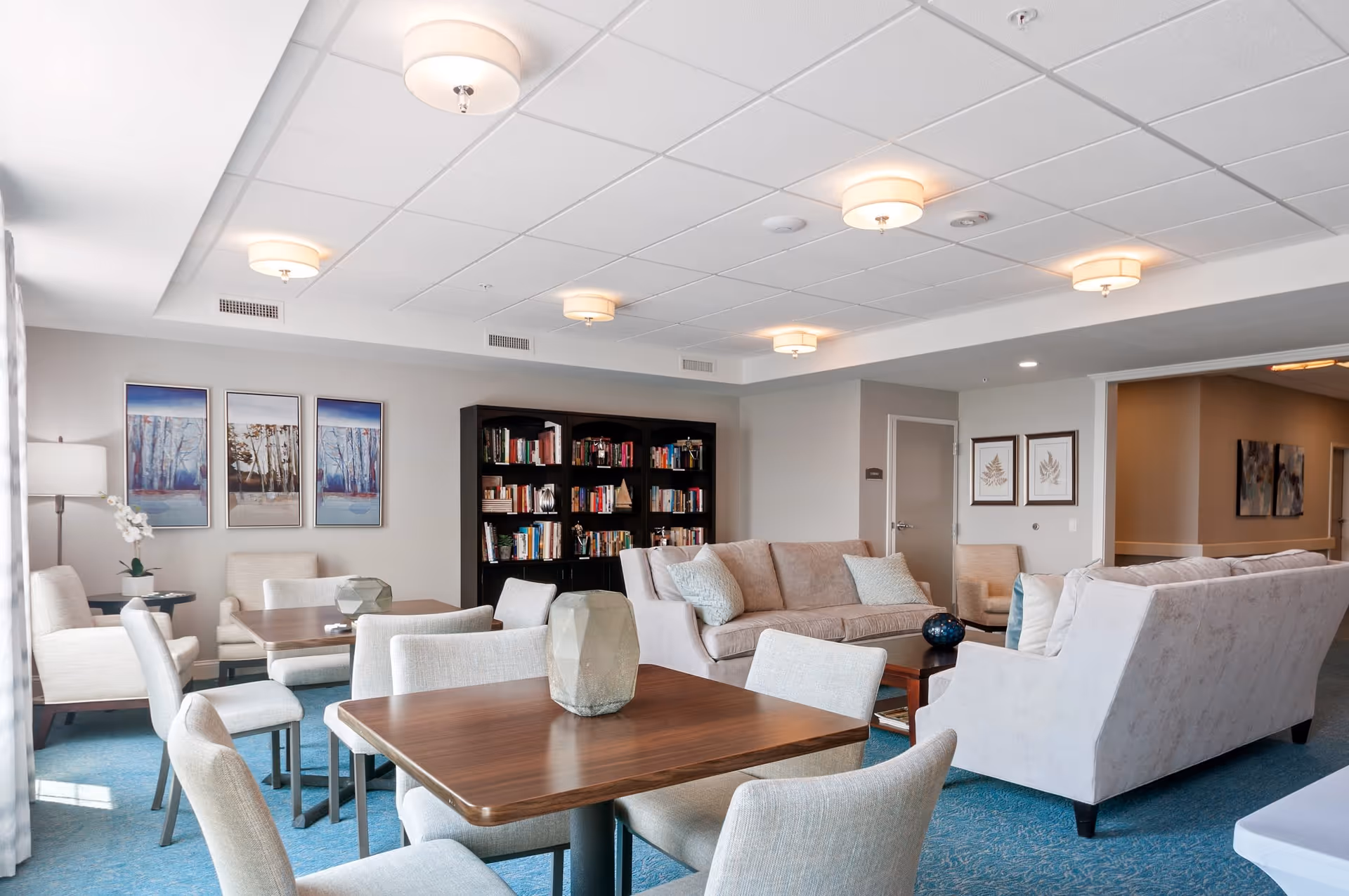 A bright and spacious common living area in a senior living facility featuring multiple beige sofas and armchairs arranged around wooden tables. There is a large bookshelf filled with books against the wall, three framed landscape paintings, and soft ceiling lights illuminating the room. The carpet is blue, and the walls are painted light gray.