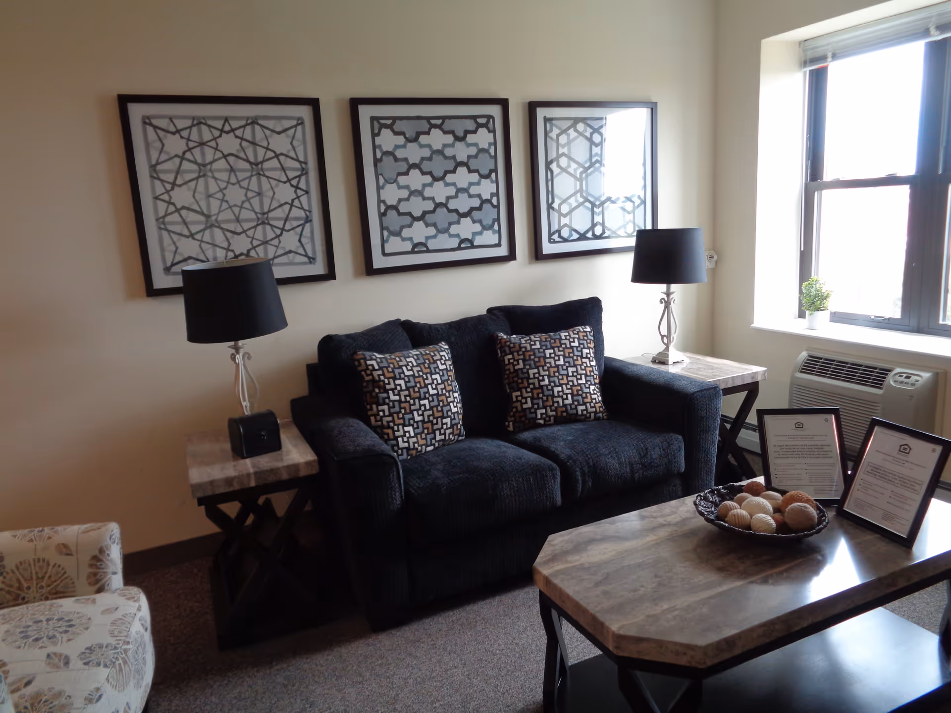 A cozy living room with a dark blue loveseat adorned with two patterned pillows, flanked by two side tables each with a black lamp. Above the loveseat are three framed geometric art pieces. A window on the right lets in natural light, with a small potted plant on the windowsill and an air conditioning unit below. In front of the loveseat is a coffee table with a decorative bowl of assorted balls and two framed documents.