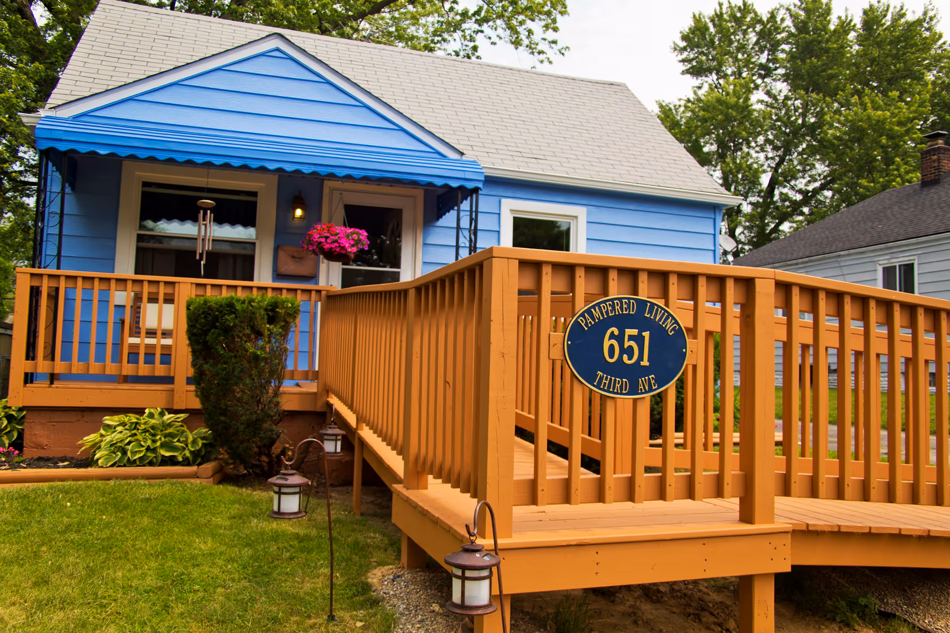 Exterior view of a small blue house with a gray roof and a wooden wheelchair ramp leading to the front door. The ramp has a blue and yellow sign that reads 'Pampered Living 651 Third Ave'. There are green bushes, a hanging flower pot with pink flowers, and small lanterns along the ramp. Trees and neighboring houses are visible in the background.
