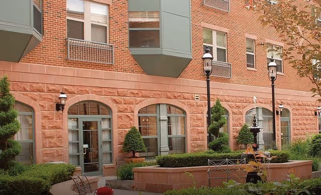 Outdoor courtyard area of a brick building with large windows and green trim. The courtyard features trimmed bushes, a small fountain, benches, and vintage-style street lamps. Trees with autumn leaves are visible on the right side.