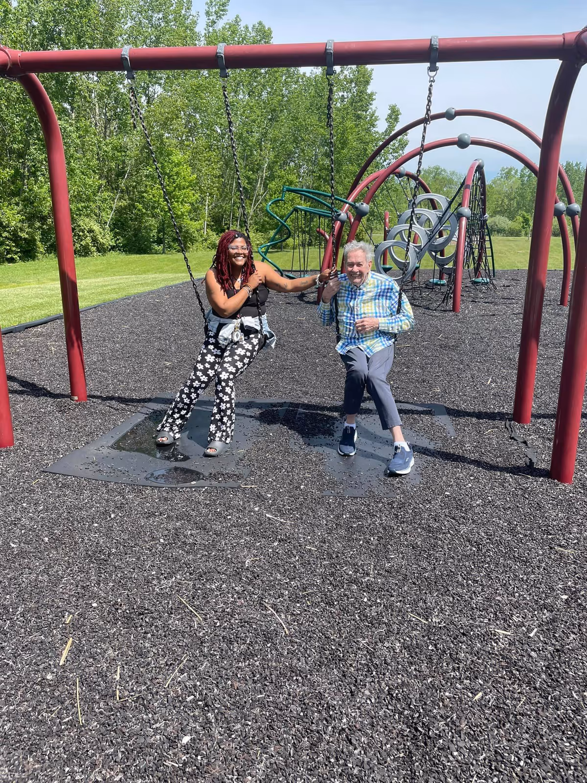 Two people sitting on swings in a playground area with rubber mulch ground covering. One person is a woman with long braided hair wearing a black tank top and floral pants, smiling and holding hands with an older man wearing a plaid shirt and gray pants. Behind them are various playground climbing structures and green trees in the background under a clear sky.