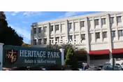 Exterior view of a multi-story building with a sign in front that reads Heritage Park Rehab & Skilled Nursing. The building has multiple windows and there are trees and parked cars visible.
