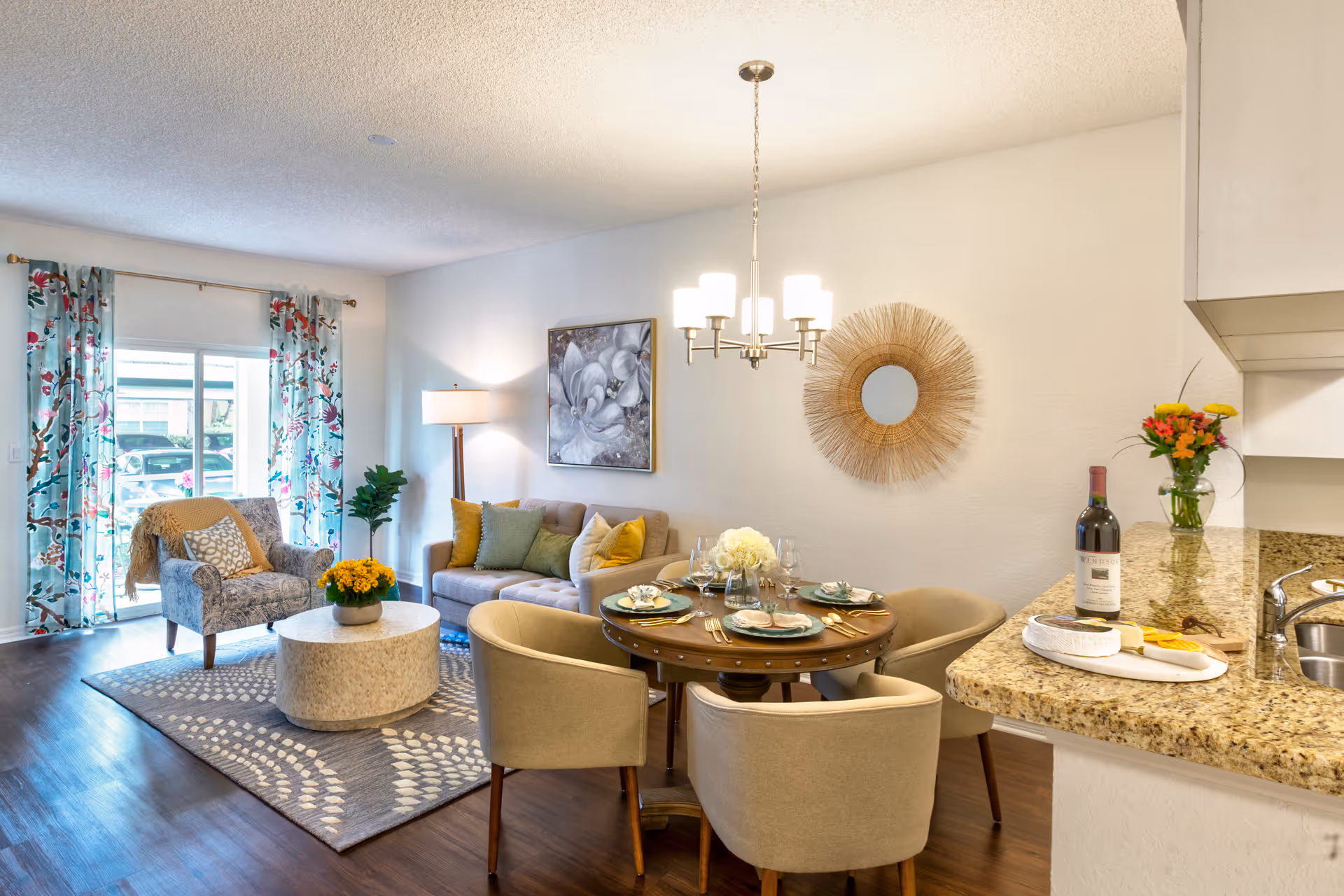 A bright and cozy living and dining area with a round dining table set for four, beige upholstered chairs, a beige sofa with green and yellow cushions, a patterned armchair, a round coffee table with a yellow flower pot, floral curtains on a sliding glass door, a floor lamp, wall art, and a kitchen counter with a bottle of wine, flowers, and a cheese platter.