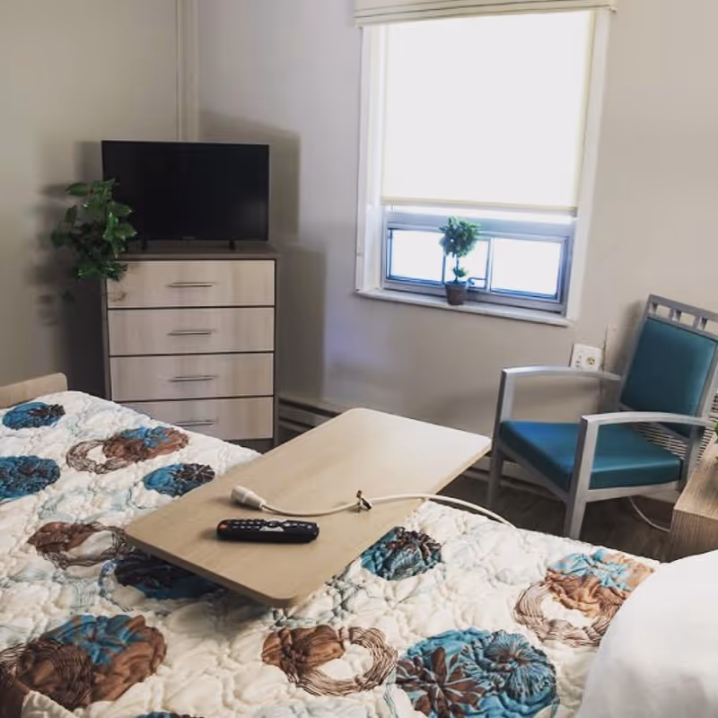 A room with a bed covered by a quilt with blue and brown circular patterns. On the bed is a wooden overbed table with a TV remote and a call button. In the background, there is a small dresser with a flat-screen TV on top and a green plant beside it. A window with a white blind partially drawn is above a blue cushioned chair with metal armrests.