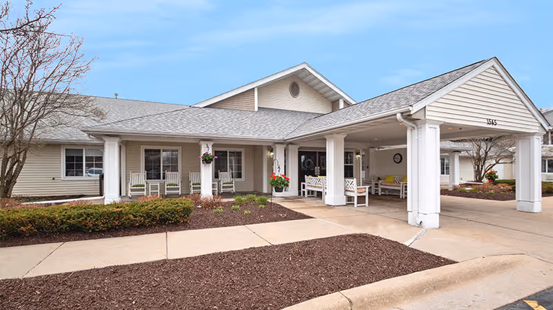 Exterior view of a single-story senior living facility building with a covered entrance supported by white columns. There are benches and hanging flower pots under the covered area, and a sidewalk leading to the entrance. The building has beige siding and a gray shingled roof, with some landscaping including bushes and small trees.