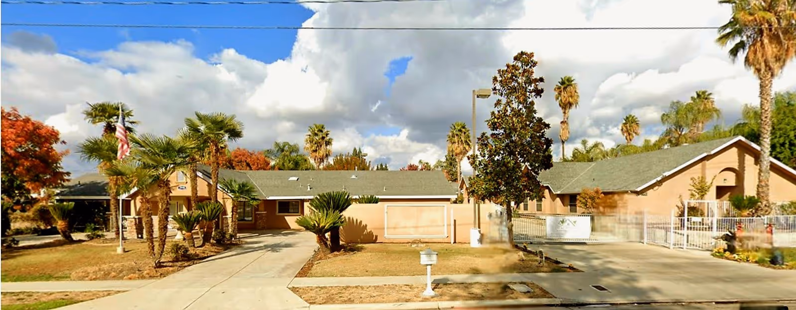 Exterior view of a single-story residential building with a green roof, surrounded by palm trees and other greenery under a partly cloudy sky. There is a driveway and a sidewalk in front of the building.