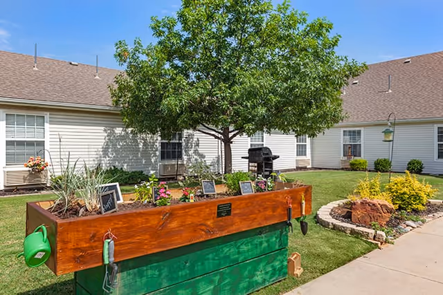 Outdoor garden area at Brookdale Richland Hills featuring a raised wooden planter box with various plants and small chalkboard labels, a green watering can hanging on the side, a tree in the background, and a barbecue grill near the building with white siding and multiple windows under a clear blue sky.
