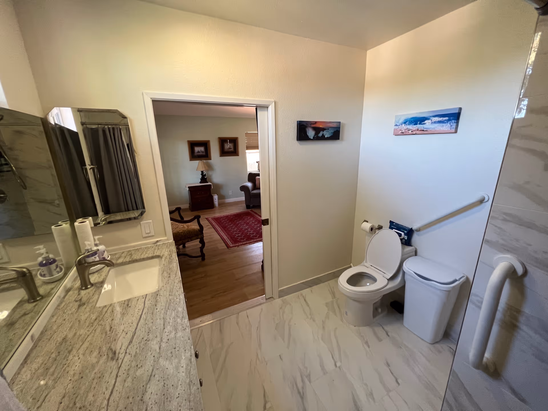 A clean and modern bathroom with a marble countertop sink on the left, a toilet with the seat up on the right, a trash bin next to the toilet, and grab bars on the wall and shower door. The bathroom has light-colored walls and a tiled floor. Through the open door, a living room area with wooden flooring, a red rug, chairs, and framed pictures on the wall is visible.