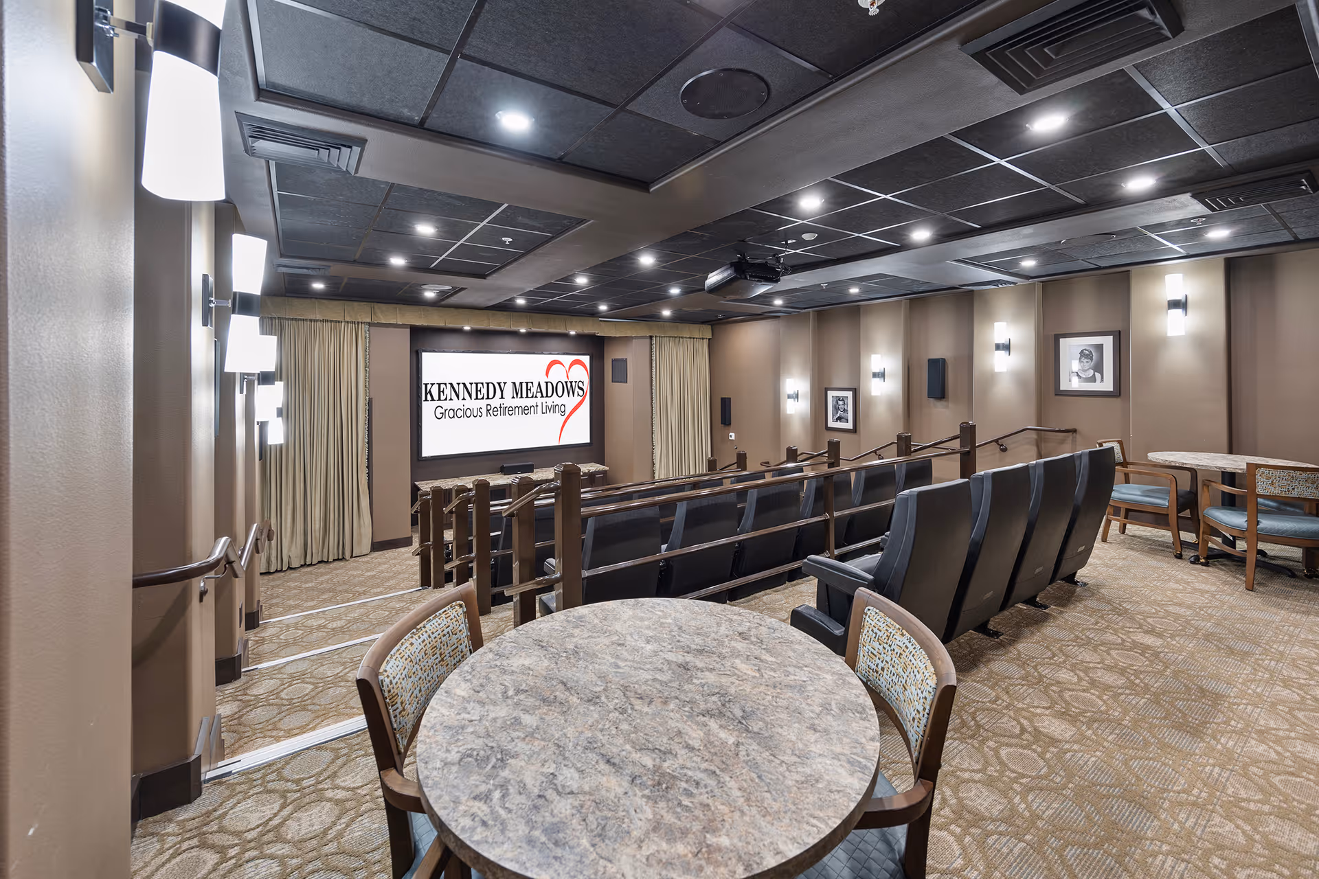 Interior view of a small theater room with tiered seating, a large screen displaying the Kennedy Meadows Gracious Retirement Living logo, round tables with chairs in the foreground, beige walls with framed pictures, and ceiling lights.