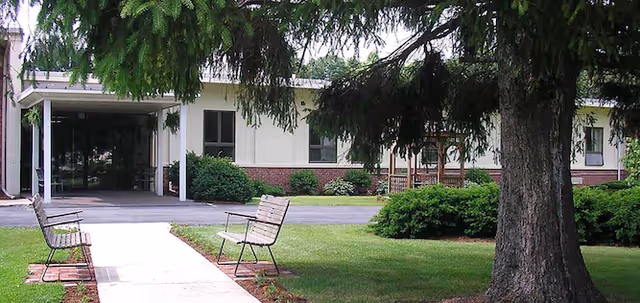 Exterior view of Michaux Manor Living Center showing a covered entrance with glass doors, a paved walkway leading to the entrance, two benches on either side of the walkway, green grass, bushes, and a large tree in the foreground.