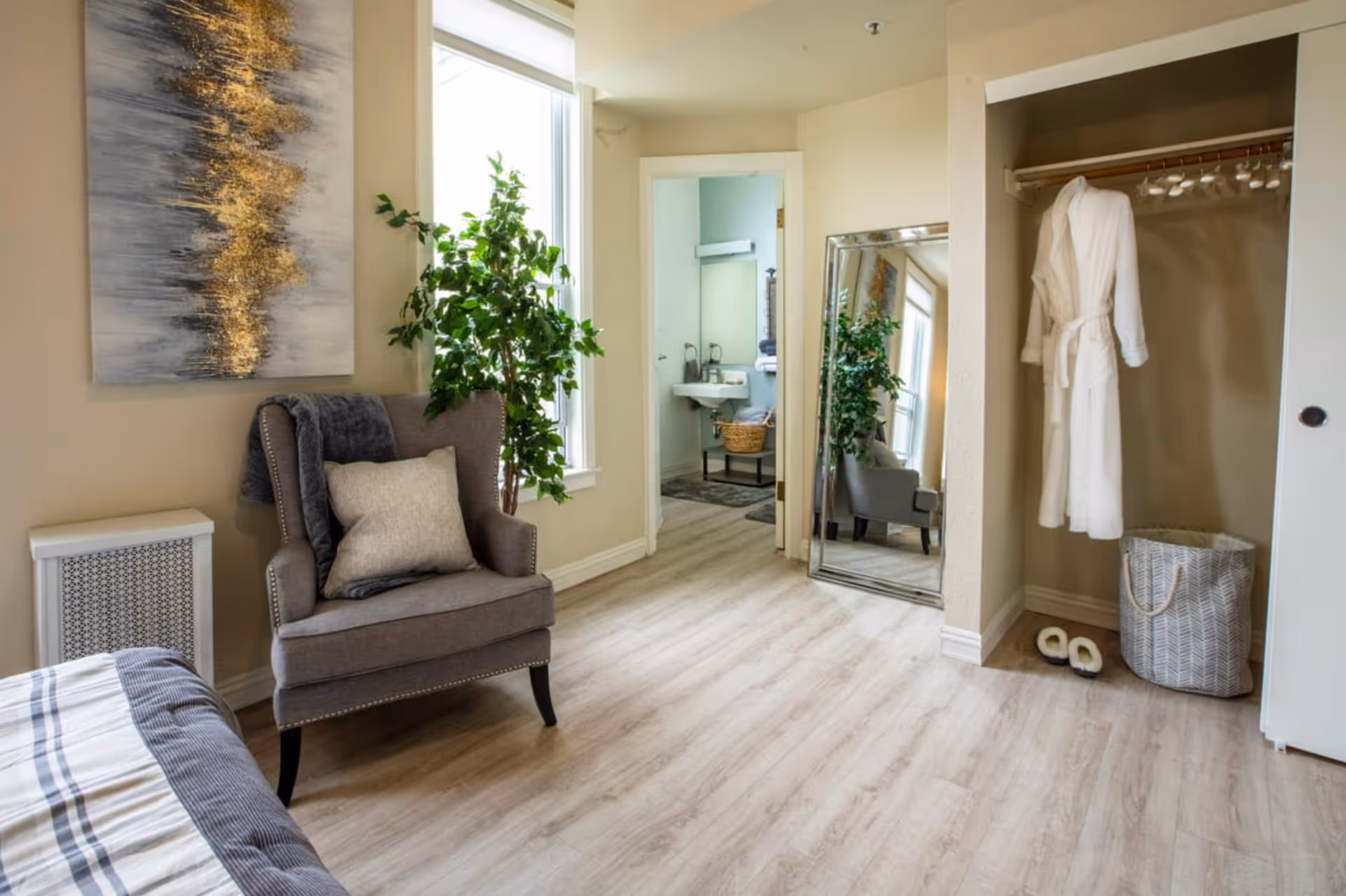 A cozy senior living bedroom featuring a gray armchair with a pillow and throw blanket, a large window with natural light, a tall green plant, a full-length mirror leaning against the wall, an open closet with a white robe hanging and slippers on the floor, and a glimpse into a small bathroom with a sink and basket.