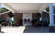 Entrance of Williamsburg Nursing Home with a covered driveway supported by white columns, brick walls, hanging plants, and seating areas near the double glass doors.