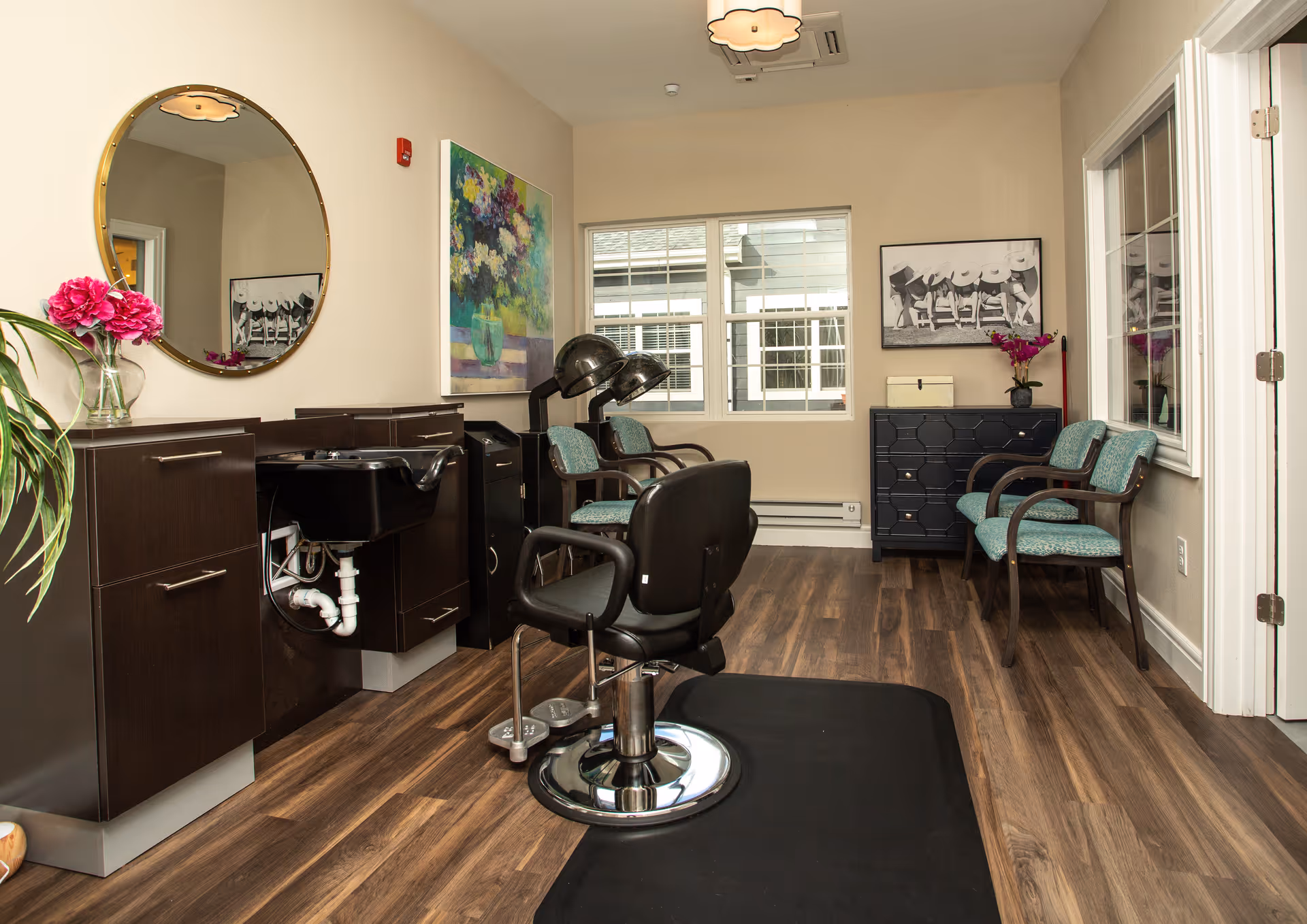 Interior view of a hair salon area in a senior living facility featuring a black salon chair on a black mat, two hair drying chairs with hooded dryers, a black sink with dark brown cabinetry, a round mirror on the wall, colorful floral artwork, two teal upholstered chairs, a black chest of drawers, and a window letting in natural light.