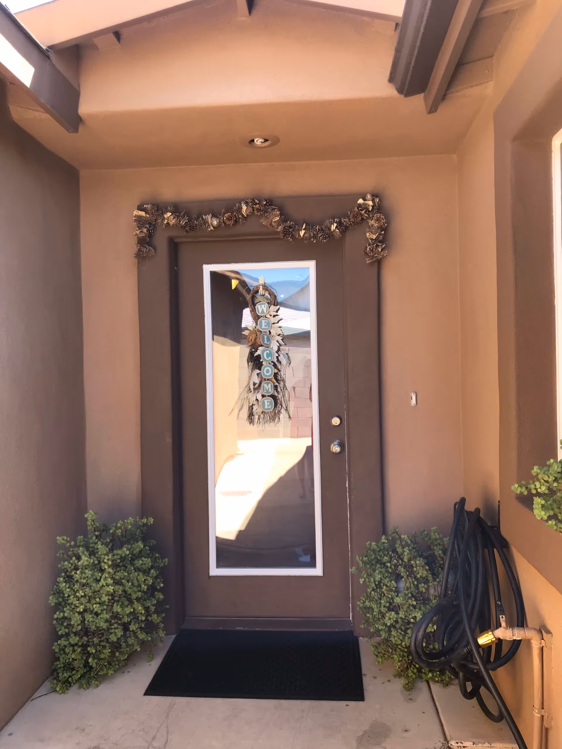 Front door entrance of a building with a glass panel door decorated with a hanging sign that says 'WELCOME'. The door is framed by brown walls and has a garland above it. There are green plants on either side of the door and a black garden hose coiled on the right side.