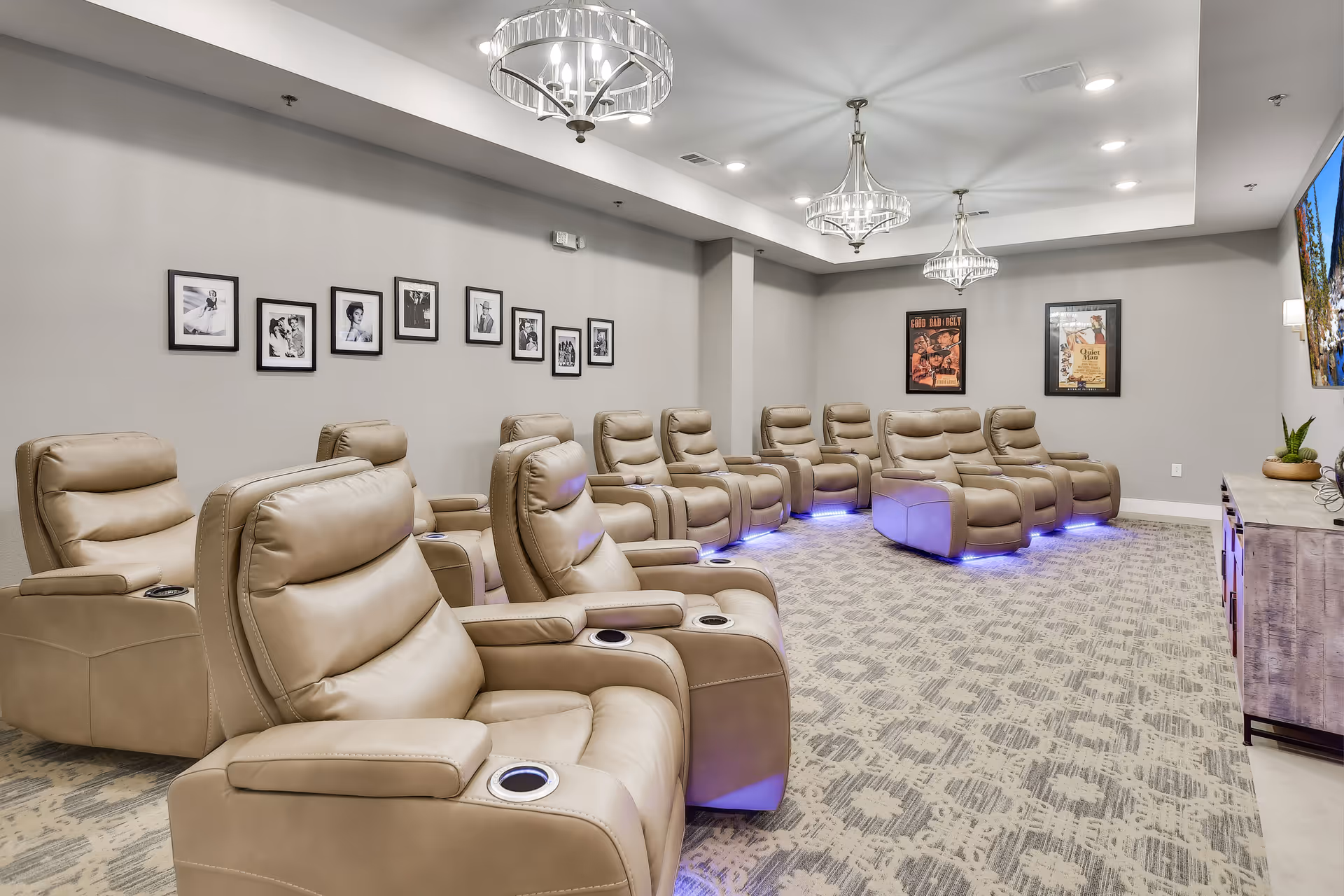 A modern media room with two rows of beige leather recliner chairs featuring cup holders. The room has a patterned carpet, gray walls adorned with framed black and white photos and movie posters, and three elegant ceiling light fixtures. A large flat-screen TV is mounted on the right wall above a wooden console with decorative plants.