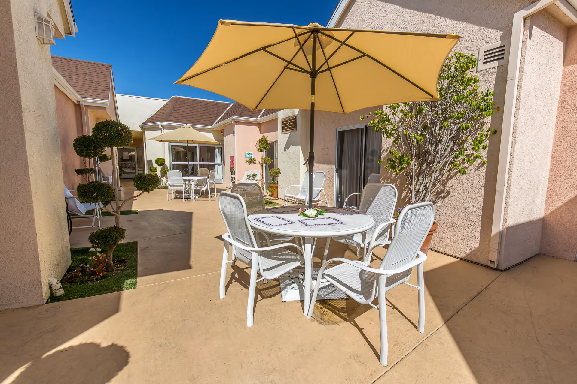 Outdoor patio area at Windsor Terrace Healthcare Center with white metal chairs and round tables shaded by large yellow umbrellas. The patio is surrounded by light pink building walls and decorated with potted plants and neatly trimmed topiary trees under a clear blue sky.