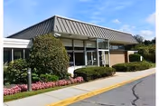 Exterior view of a single-story building with large windows, a metal roof, and landscaped bushes and flowers along the sidewalk under a blue sky with some clouds.