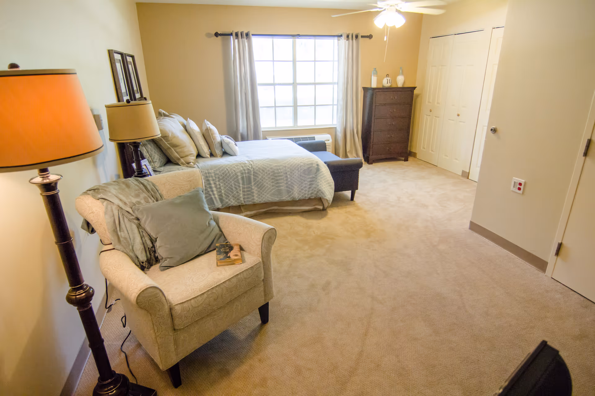 A cozy bedroom with a large window covered by light curtains. The room features a bed with multiple pillows, a cushioned armchair with a throw blanket and a book on it, a tall floor lamp, a dark wooden dresser with decorative vases on top, and a ceiling fan with a light. The walls are painted beige and the floor is carpeted.