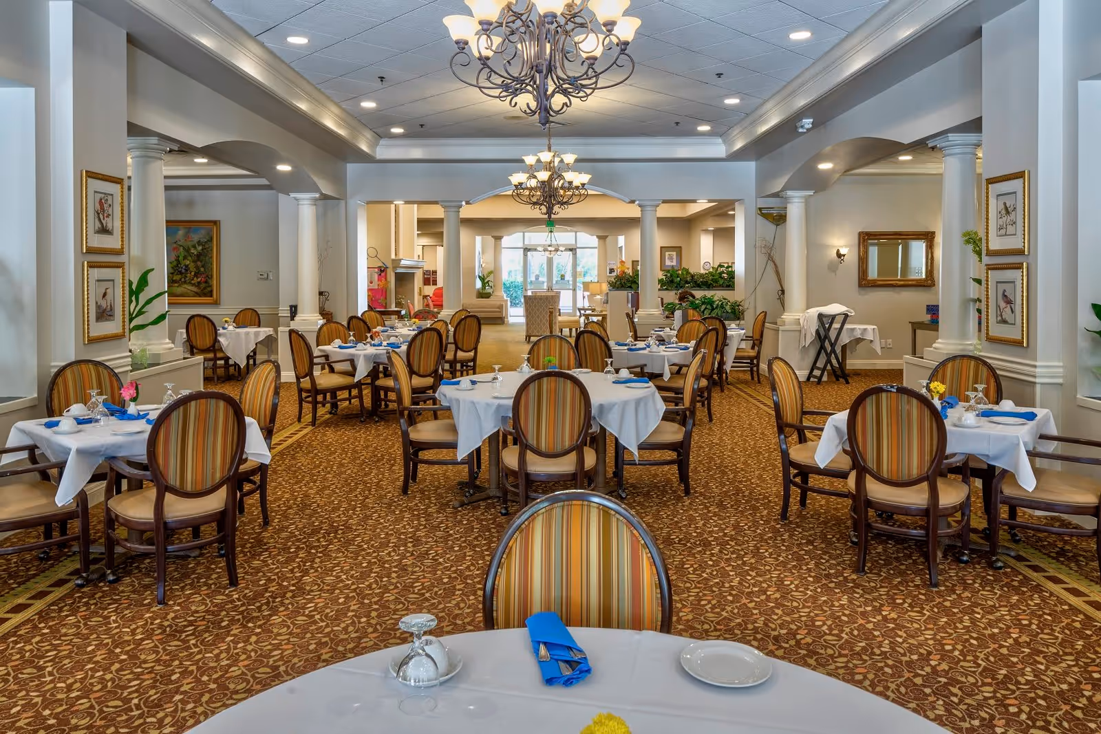 A spacious and elegant dining room with multiple round tables covered with white tablecloths, each set with plates, glasses, and blue napkins. The room features striped upholstered chairs, ornate chandeliers hanging from the ceiling, decorative columns, framed artwork on the walls, and a patterned carpet. The far end of the room opens to a seating area with large windows letting in natural light.