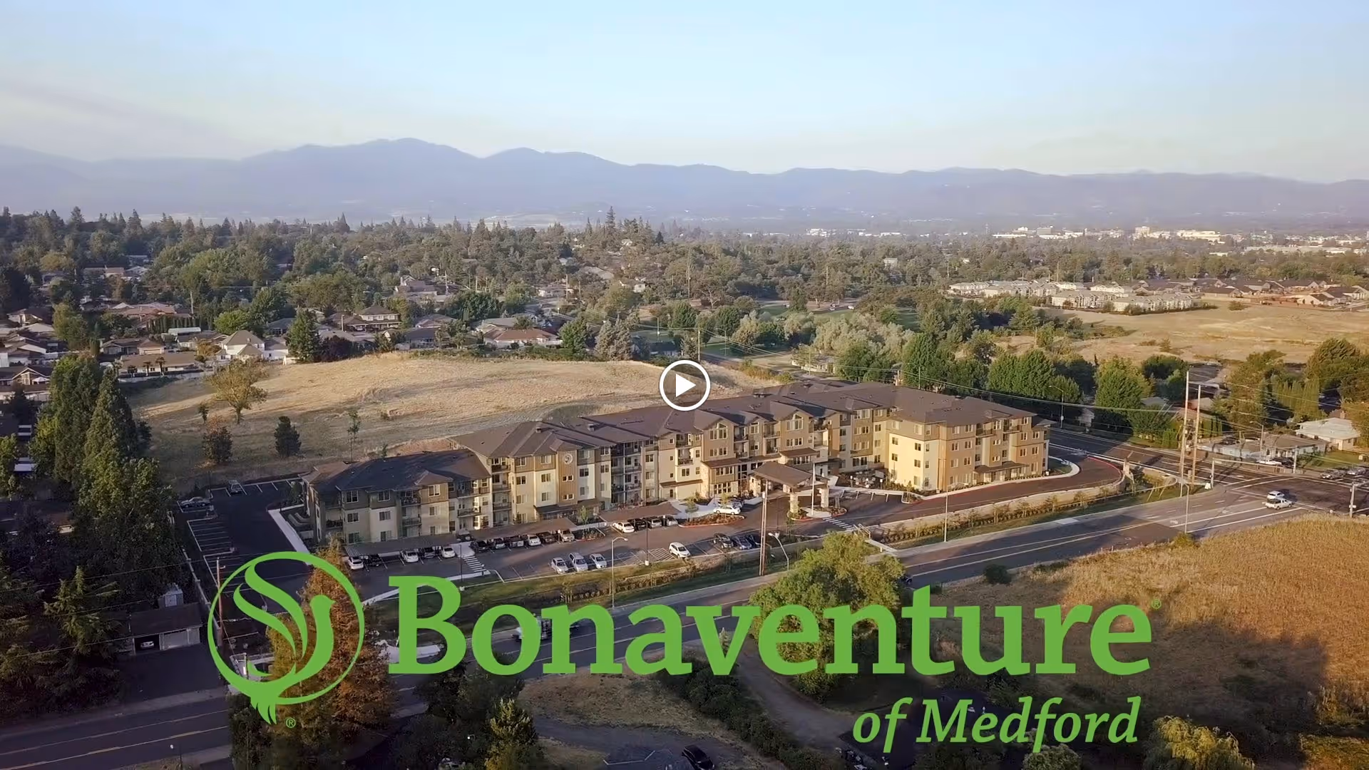 Aerial view of Bonaventure of Medford, a large multi-story senior living facility surrounded by trees, roads, and open fields with mountains visible in the distance under a clear sky.
