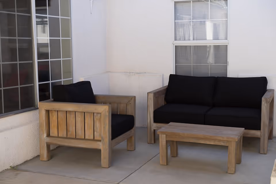 Outdoor seating area with wooden furniture including a black cushioned armchair, a black cushioned sofa, and a wooden coffee table, set against a white wall with two windows.