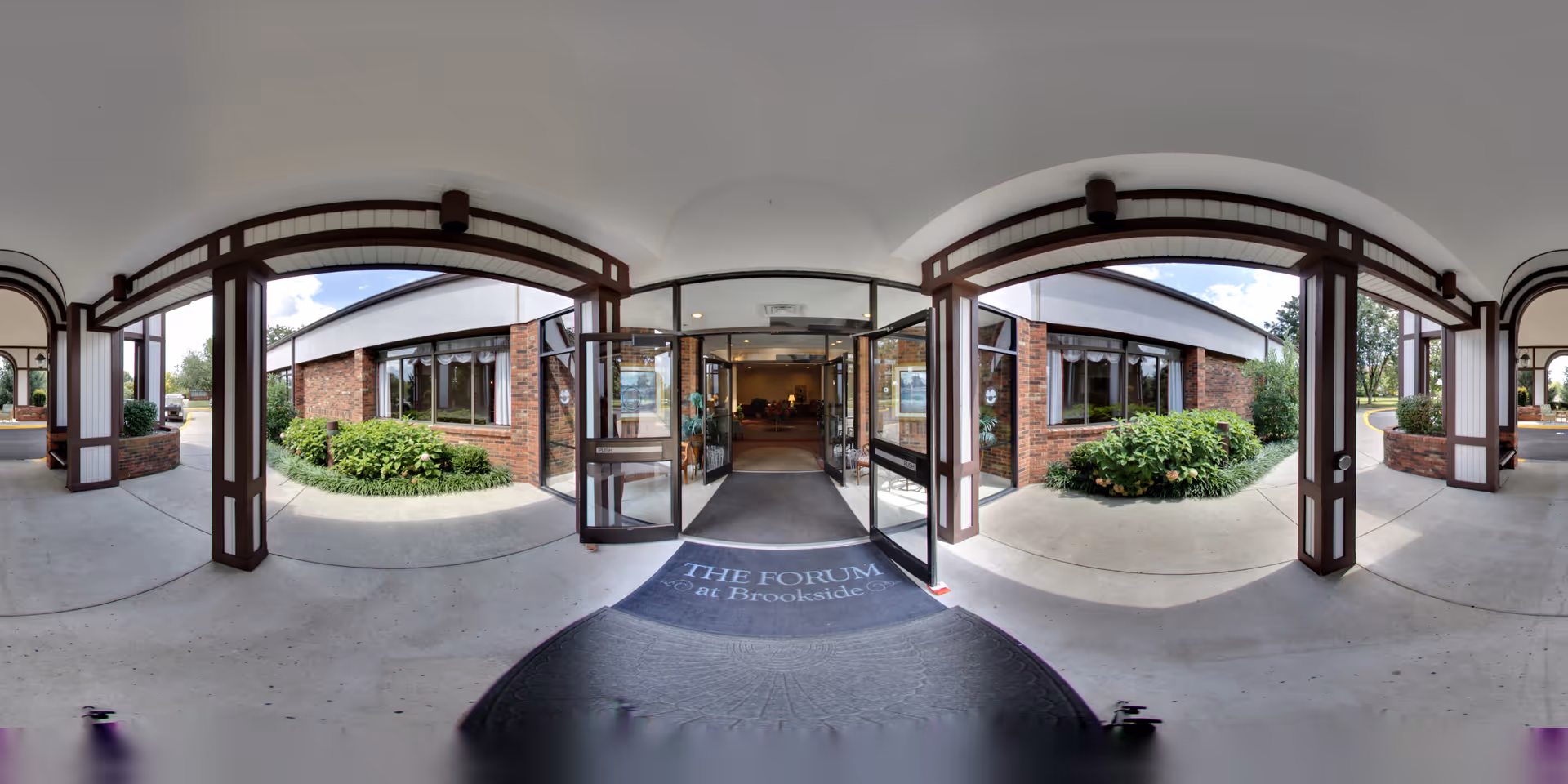 Entrance area of The Forum at Brookside senior living facility showing a covered walkway with wooden beams and brick walls, glass double doors leading inside, and greenery on either side of the entrance.