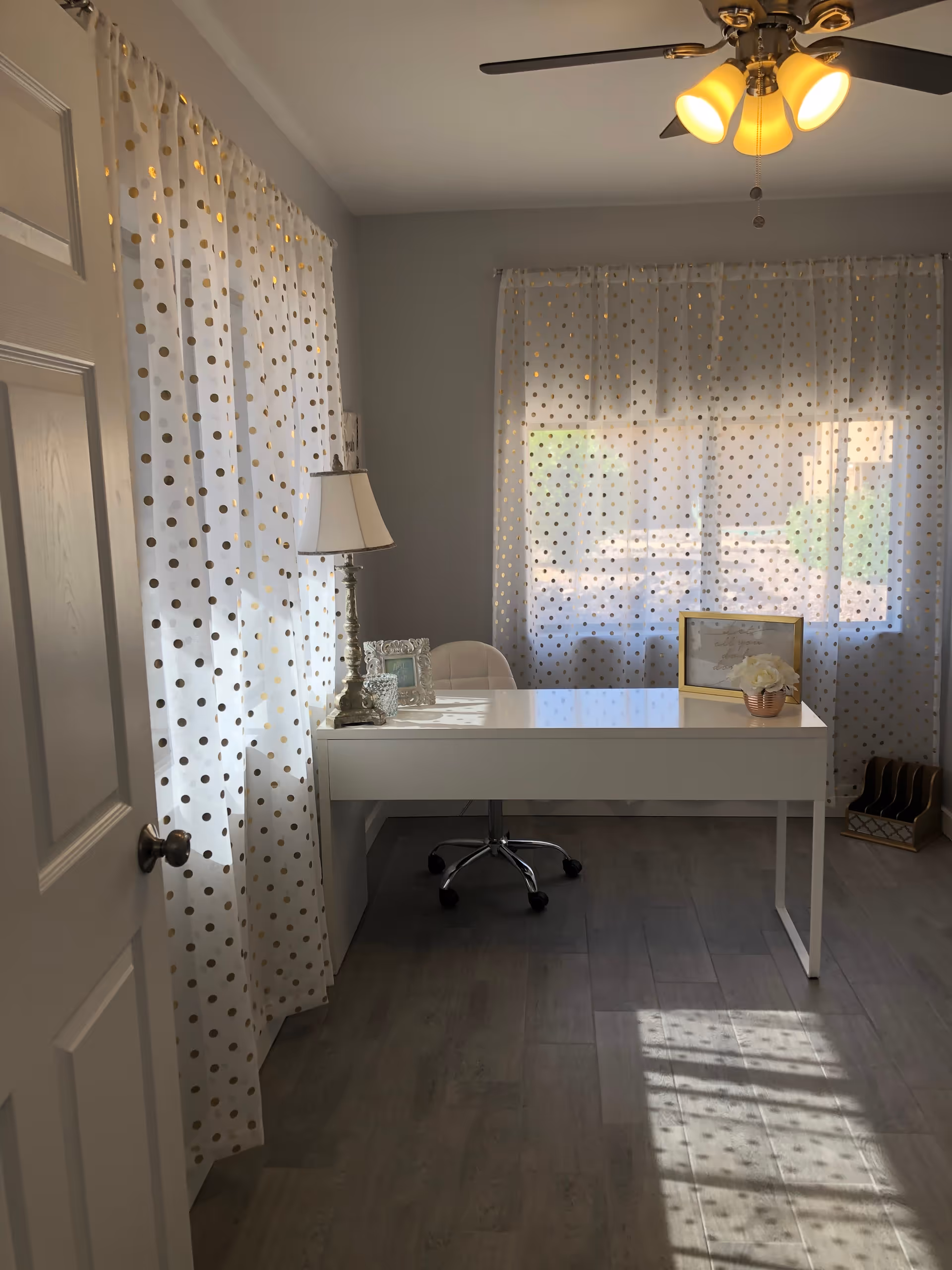 A bright room with sheer white curtains featuring gold polka dots on two windows. A white desk with a white swivel chair is positioned in front of one window. On the desk are a decorative lamp, a framed photo, a framed quote, and a small flower arrangement. The floor is wood, and a ceiling fan with lights is visible above.