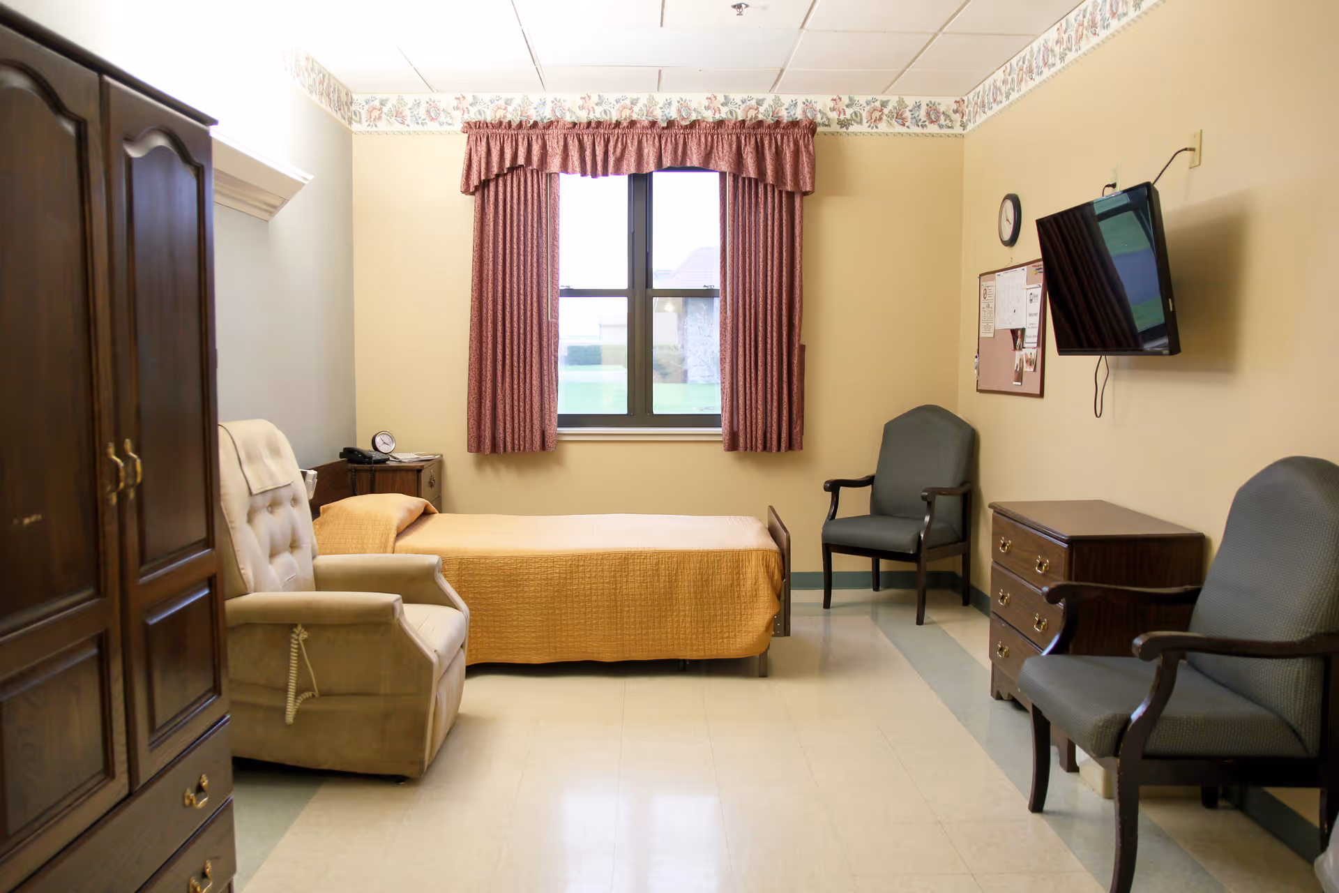 A simple, clean bedroom in a senior living facility with a single bed covered in a yellow bedspread, a beige recliner chair, two dark green armchairs, a wooden dresser, a wall-mounted TV, a clock, and a window with red curtains letting in natural light.