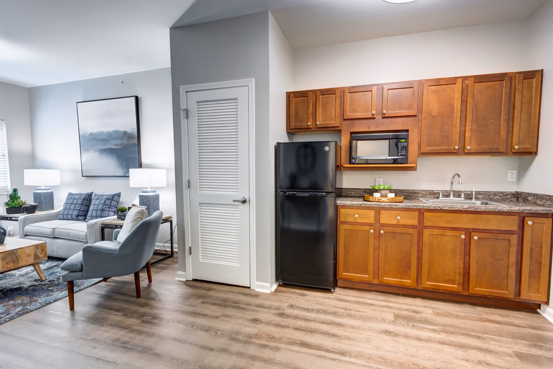 A modern living space featuring a small kitchen area with wooden cabinets, a black refrigerator, a microwave, and a sink. Adjacent to the kitchen is a cozy living room with a gray sofa, a blue armchair, two table lamps, a wooden coffee table, and a large framed artwork on the wall. The floor is covered with light wood laminate.