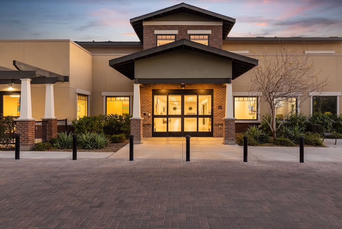 Front entrance of BridgeWater Assisted Living facility at dusk, featuring a covered entryway with brick pillars, glass double doors, and surrounding landscaping with bushes and a leafless tree.