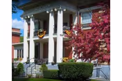 Front exterior view of a brick building with large white columns decorated with autumn wreaths, surrounded by green bushes and a tree with pink blossoms under a blue sky.